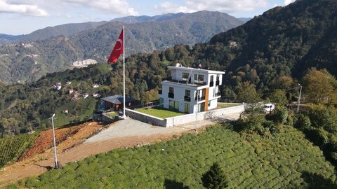 tepe platosunda çim teras ve büyük türk bayrağı direği olan çok katlı villa; çevrede dağ manzarası ve çay bahçeleri (drone çekimi, yan/arka açı)