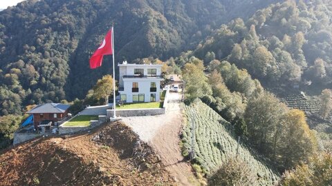 önden görünüm — geniş çim alan, ön solda türk bayrağı direği, arkada çok katlı villa; sağda çay terasları ve derin vadi manzarası