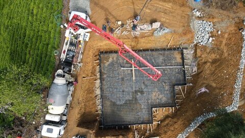 üstten görünüm — temelde radye donatısı ve kırmızı beton pompası; solda transmikser ve şantiye araçları, sağda kazı sahası
