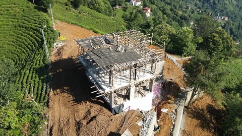 drone açı — dik yamacın üzerinde betonarme karkas ve üst döşeme ahşap kalıbı; çevrede çay terasları ve şantiye yolları