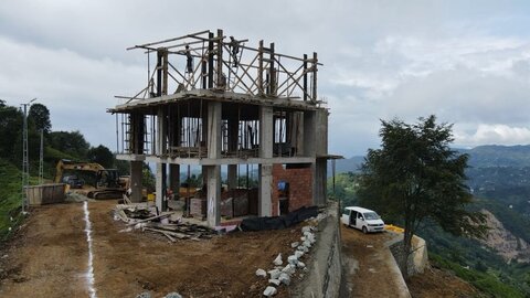 iki katlı betonarme karkas; üst katta ahşap kalıp/iskelet, açık kolon-kirişler; solda iş makineleri, sağda platformda beyaz araç ve vadi manzarası