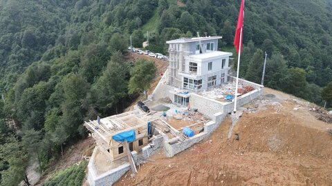 Drone ile tepe üstü genel görünüm — çok katlı ana kütle, alt kottaki ek yapı ve büyük Türk bayrağı
